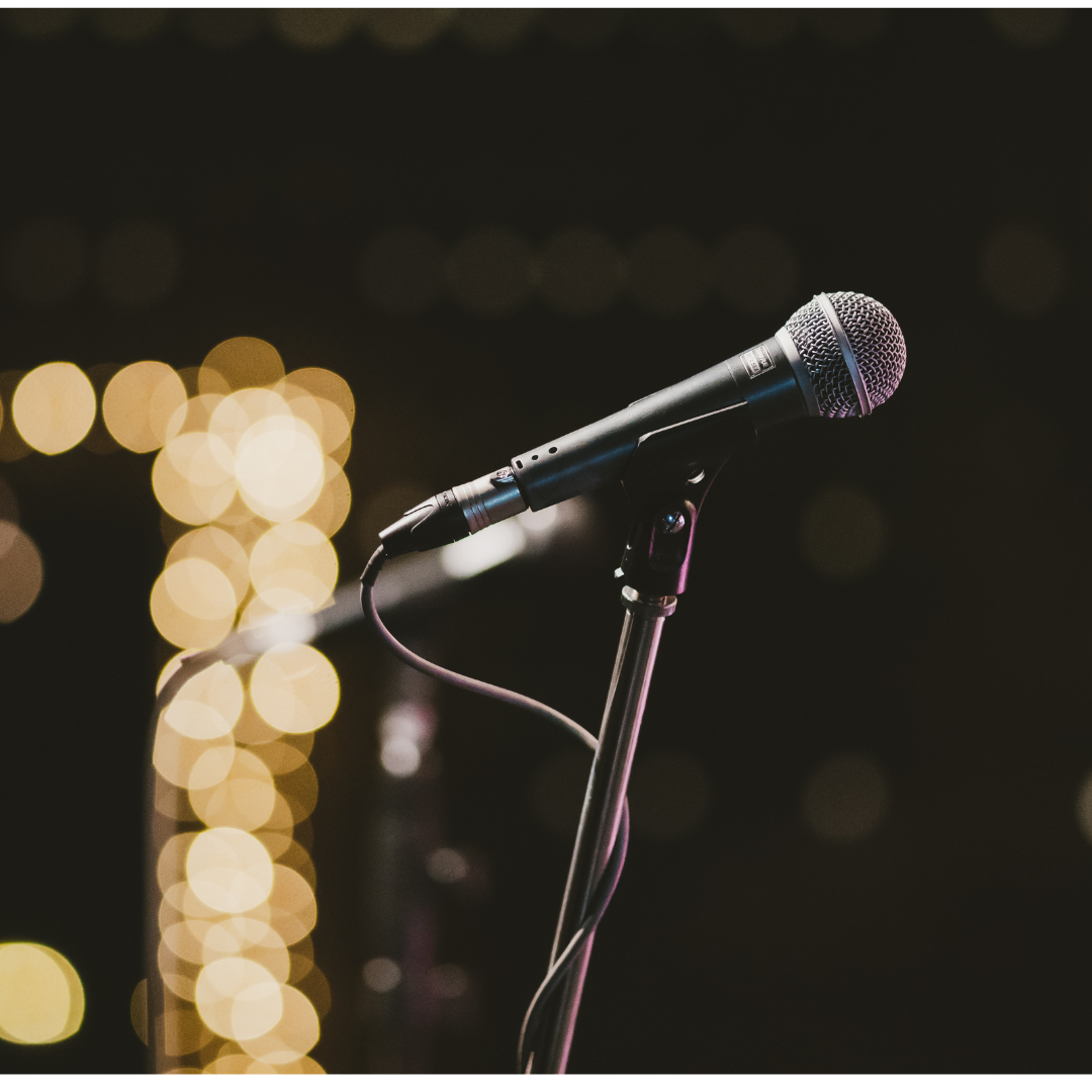 A closeup of a stage microphone on a black background with blurred lights.