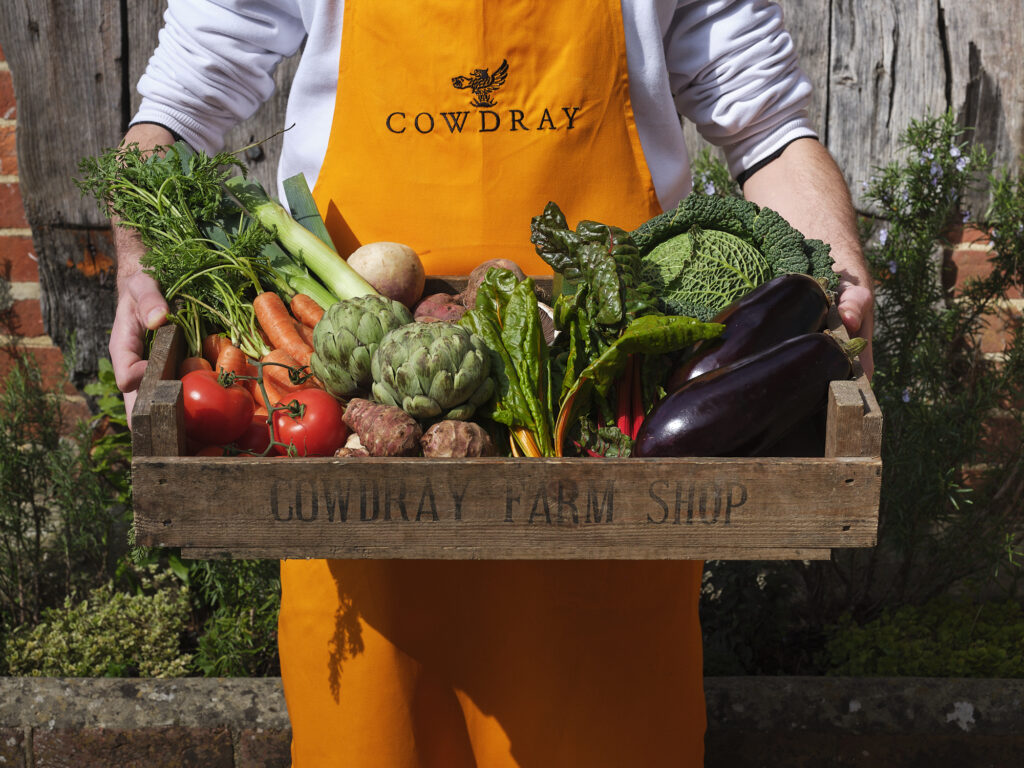 (c) Cowdray Estate – 220322_0372 A closeup of a Cowdray Farm Shop staff member wearing a yellow Cowdray apron holding a wooden box full of fresh seasonal vegetables in Midhurst, West Sussex.