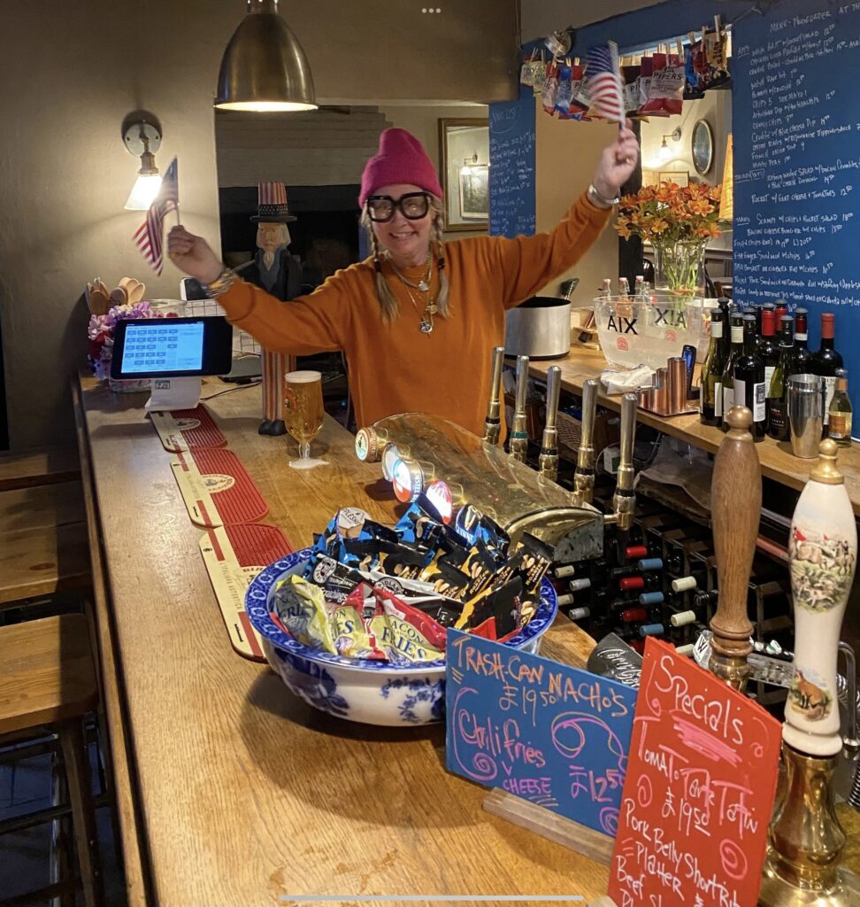 (c) The Hollist Arms – Screenshot 1 The bar counter of The Hollist Arms pub in West Sussex, with a staff member waving American flags.