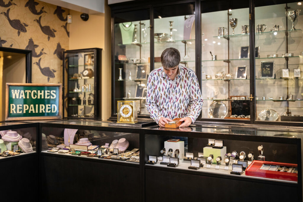 (c) Christopher Ison Photography – ISON_200715_Allnutt_549998 Inside J E Allnutt & Son Ltd shopfront in West Street, Midhurst with a shopkeeper behind the counter which shows vintage watches, jewellery and silver.