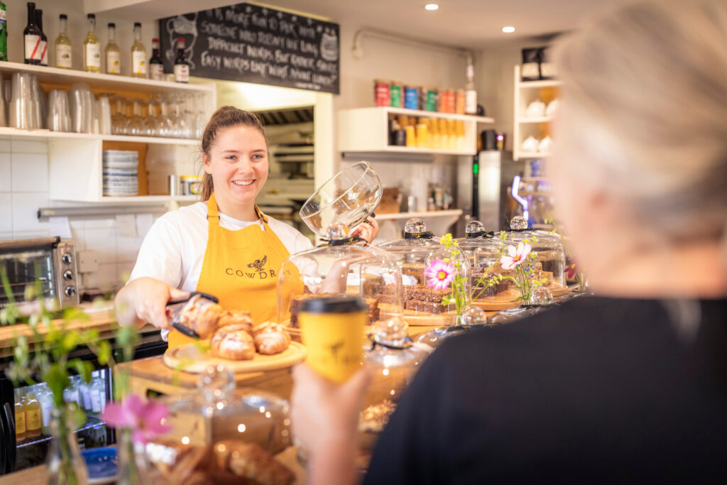 Staff member of Cowdrey Farm Shop & Cafe wearing yellow Cowdrey apron behind the counter serving a customer a pastry.