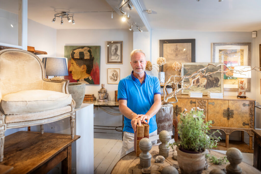 (c) Christopher Ison Photography – ISON_230624_OliveInt_541414 Inside of Olive Interiors in Midhurst, West Sussex with the shopkeeper surrounded by country house style furniture and antiques.