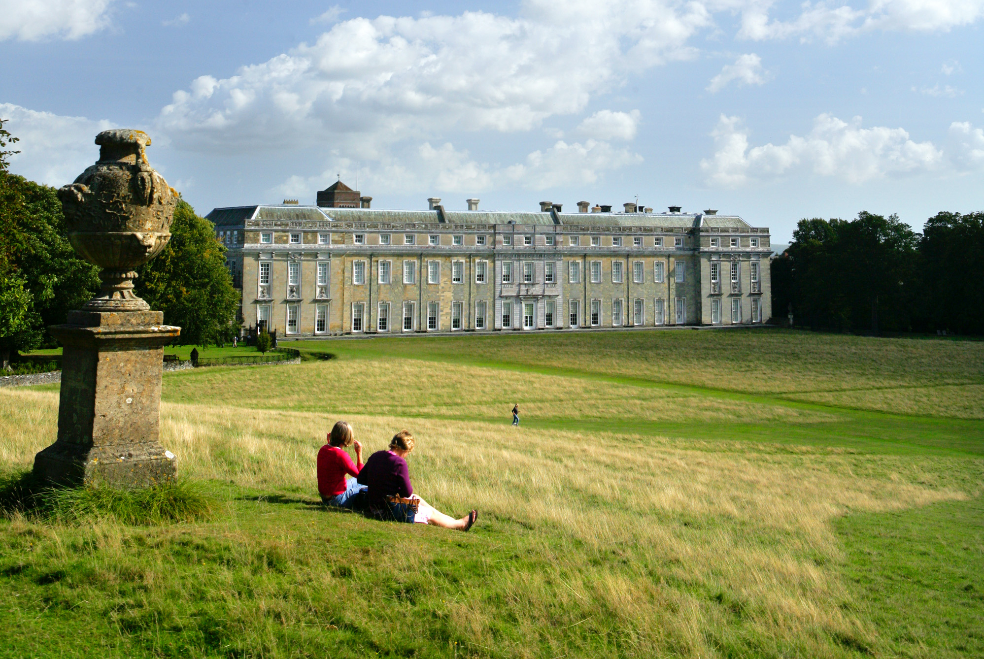 A view of Petworth House in Petworth Park surrounded by green fields and walkers.