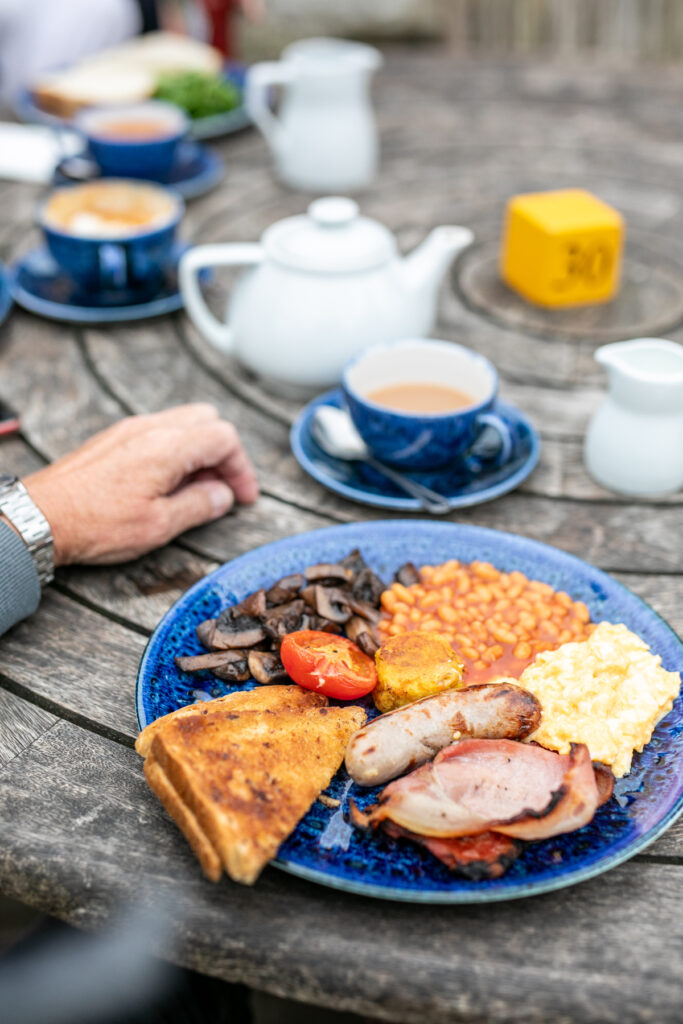 (c) Cowdray Estate – Cowdray-Estate-George-Gunn-8343 Closeup of an English breakfast and cups of tea made by Cowdray Farm Shop & Cafe, on an outdoor wooden table.